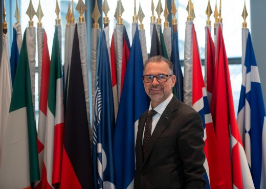 Aschbacher, lächelnd und Brille-Tragend, steht vor einer Wand voller Flaggen der ESA Mitgliedsländer