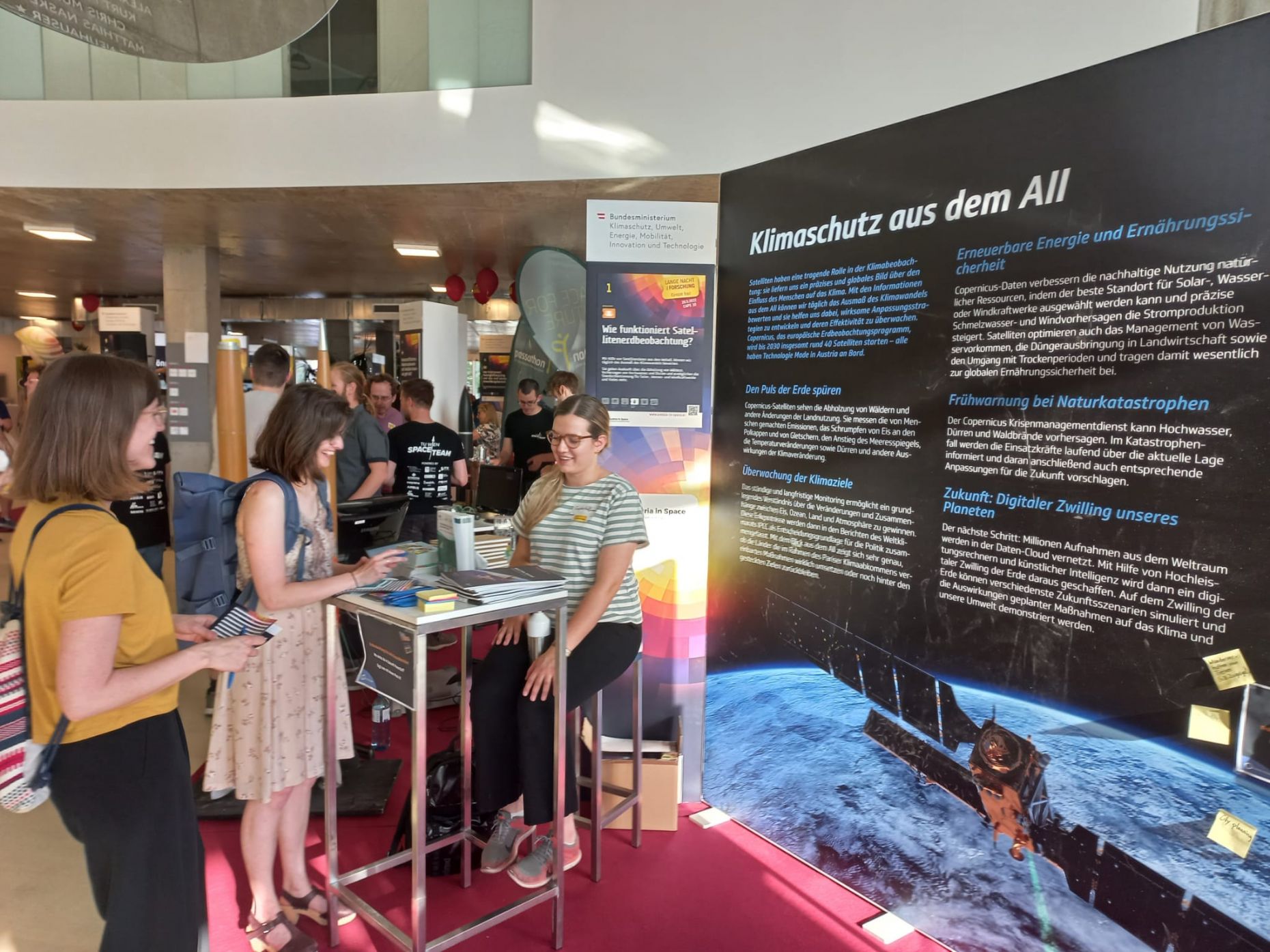 Ein Messestand, der über Erdbeobachtung und Weltraumaktivitäten in Österreich informiert. Davor stehen interessierte Besucher:innen, die aufmerksam die ausgestellten Plakate studieren.