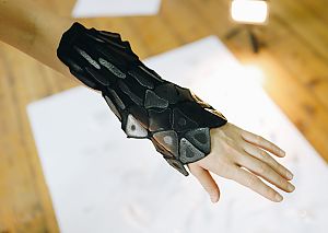 Eine Nahaufnahme eines Arms mit einer schwarzen, strukturierten Manschette aus flexiblen, geometrischen Elementen. Das Design erinnert an eine futuristische oder biomimetische Rüstung.