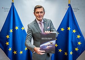Der Minister blickt in die Kamera und hält die Druckfassung der österreischischen Weltraumstrategie in die Kamera. Er trägt einen grauen Anzug und eine rosa Krawatte und steht zwischen zwei blauen EU Flaggen mit den gelben, kreisförmig angeordneten Sternen und im Hintergrund ist das Logo des Bundesministeriums für Innovation, Mobilität und Infrastruktur erkennbar.