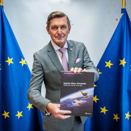 Der Minister blickt in die Kamera und hält die Druckfassung der österreischischen Weltraumstrategie in die Kamera. Er trägt einen grauen Anzug und eine rosa Krawatte und steht zwischen zwei blauen EU Flaggen mit den gelben, kreisförmig angeordneten Sternen und im Hintergrund ist das Logo des Bundesministeriums für Innovation, Mobilität und Infrastruktur erkennbar.