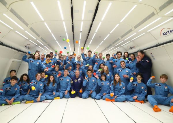 Gruppenfoto der strahlenden Teilnehmer:innen an Astronaut for a day. Man sieht ca. 30 jugendliche Mädchen und Jungen in hellblauen Overalls und neonfarbenen Socken, die bunte kleine Bälle werfen und vier erwachsene Betreuer in dunkelblauen Overalls. 
