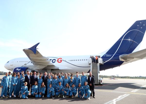 Gruppenfoto der Teilnehmer:innen an Astronaut for a day auf einer Landebahn vor einem weißen Flugzeug. Die Jugendlichen tragen hellblaue Overalls, die Erwachsenen tragen dunkle Anzüge.