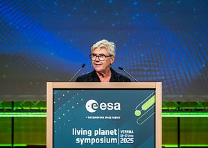 Eine freundlich blickende Frau mit weißen kurzen Haaren und schwarzerm runder Brille spricht auf einem Podium in zwei Mikrofone.