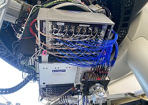 Mehrere elektronische Module und Steuergeräte sind mit zahlreichen blauen und braunen Kabeln verbunden. Die Komponenten tragen Aufschriften wie ‚General Atomics‘ und ‚qtlabs‘. Das System ist an einer mechanischen Struktur befestigt, umgeben von Kabeltrassen und Technikgehäusen.