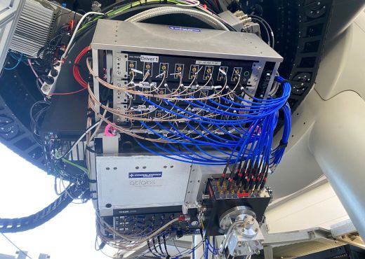 Mehrere elektronische Module und Steuergeräte sind mit zahlreichen blauen und braunen Kabeln verbunden. Die Komponenten tragen Aufschriften wie ‚General Atomics‘ und ‚qtlabs‘. Das System ist an einer mechanischen Struktur befestigt, umgeben von Kabeltrassen und Technikgehäusen.