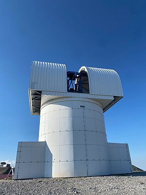Rundes, weißes Observatoriumsgebäude mit geöffneter Kuppel, durch die das Teleskop sichtbar ist. Der Himmel darüber ist klar und blau.