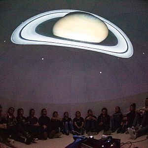 Kinder sitzenin einem abgedunkelten Raum am Boden über ihnen ist der Saturn auf die Decke projiziert.
