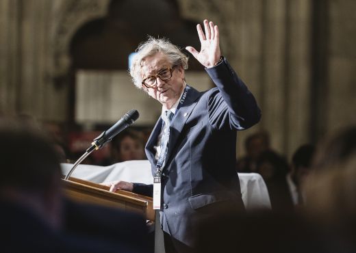 Ludwig Möller steht an einem Rednerpullt und winkt in die Kamera und in das Publikum.