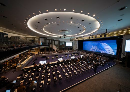 Ein großer Saal mit Tischen und einer großen Leinwand. Personen sitzen an einigen Tischen und hören den Vortragenden zu.