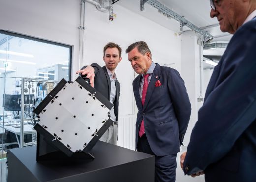 Zwei Männer in Anzügen, einer davon Innovationsminister Peter Hanke, begutachten in einem Labor einen würfelförmigen Satellitenprototyp auf einem Sockel.