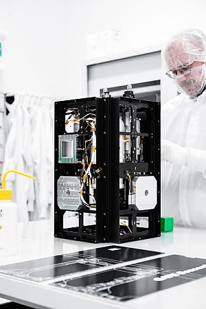 Techniker in Reinraumkleidung arbeitet an einem offenen, würfelförmigen Satellitenmodul auf einem Labortisch.