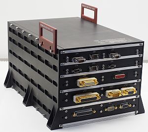 Ein schwarzes, rechteckiges Raumfahrt-Elektronikmodul mit mehreren Steckverbindern auf der Vorderseite und zwei Griffen mit den Aufschriften „Remove Before Test“ und „Remove Before Flight“.