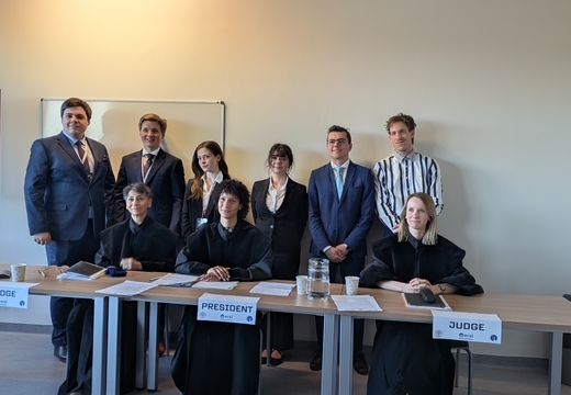 Gruppe von Teilnehmer:innen in formeller Kleidung hinter mehreren Tischen mit den Beschriftungen „Judge“ und „President“ bei einem Moot Court.