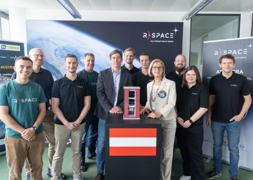 Ten R-Space employees are standing with Johanna Mikl-Leitner, Governor of Lower Austria, in front of dark blue R-Space posters. In the centre, between the people, stands a black table on which a model of their red AT-Astra satellite and a red-white-red Austrian flag can be seen.