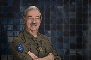 Porträt von Friedrich Teichmann in olivgrüner Uniform mit verschränkten Armen vor einem dunkelblauen Hintergrund.