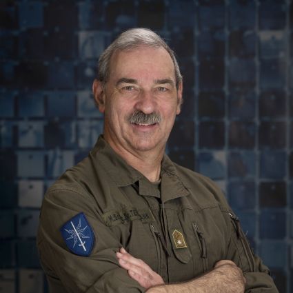 Porträt von Friedrich Teichmann in olivgrüner Uniform mit verschränkten Armen vor einem dunkelblauen Hintergrund.